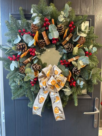 Robin’s Woodland - Bring the beauty of a traditional countryside Christmas to your home with the Robin’s Woodland Christmas Wreath. Handcrafted with fresh evergreen foliage, this natural design is beautifully decorated with aromatic cinnamon bundles, dried orange slices, rustic pine cones, vibrant red berries, soft eucalyptus foliage, and finished with a charming robin-patterned ribbon. Blending woodland textures with classic festive colours, this wreath creates a warm, welcoming display that captures the magic of the season.